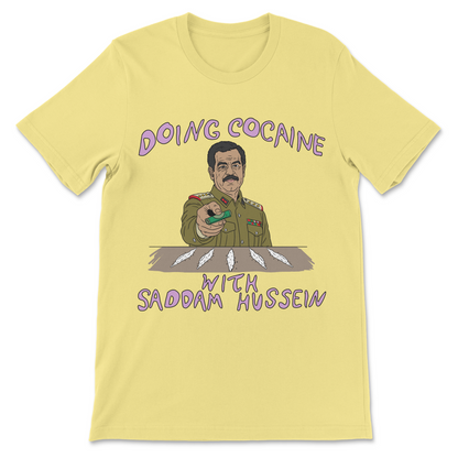 A pale yellow short-sleeve t-shirt with a graphic print of a cartoon military figure and the text 'DOING COCAINE WITH SADDAM HUSSEIN' in purple lettering.