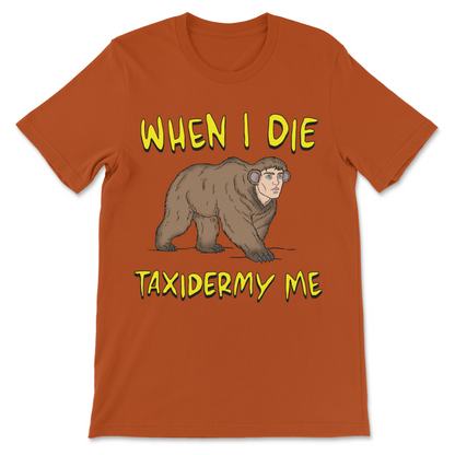 An orange crewneck t-shirt featuring a cartoon illustration of a brown bear wearing headphones, with the text 'WHEN I DIE' above and 'TAXIDERMY ME' below in a yellow font.